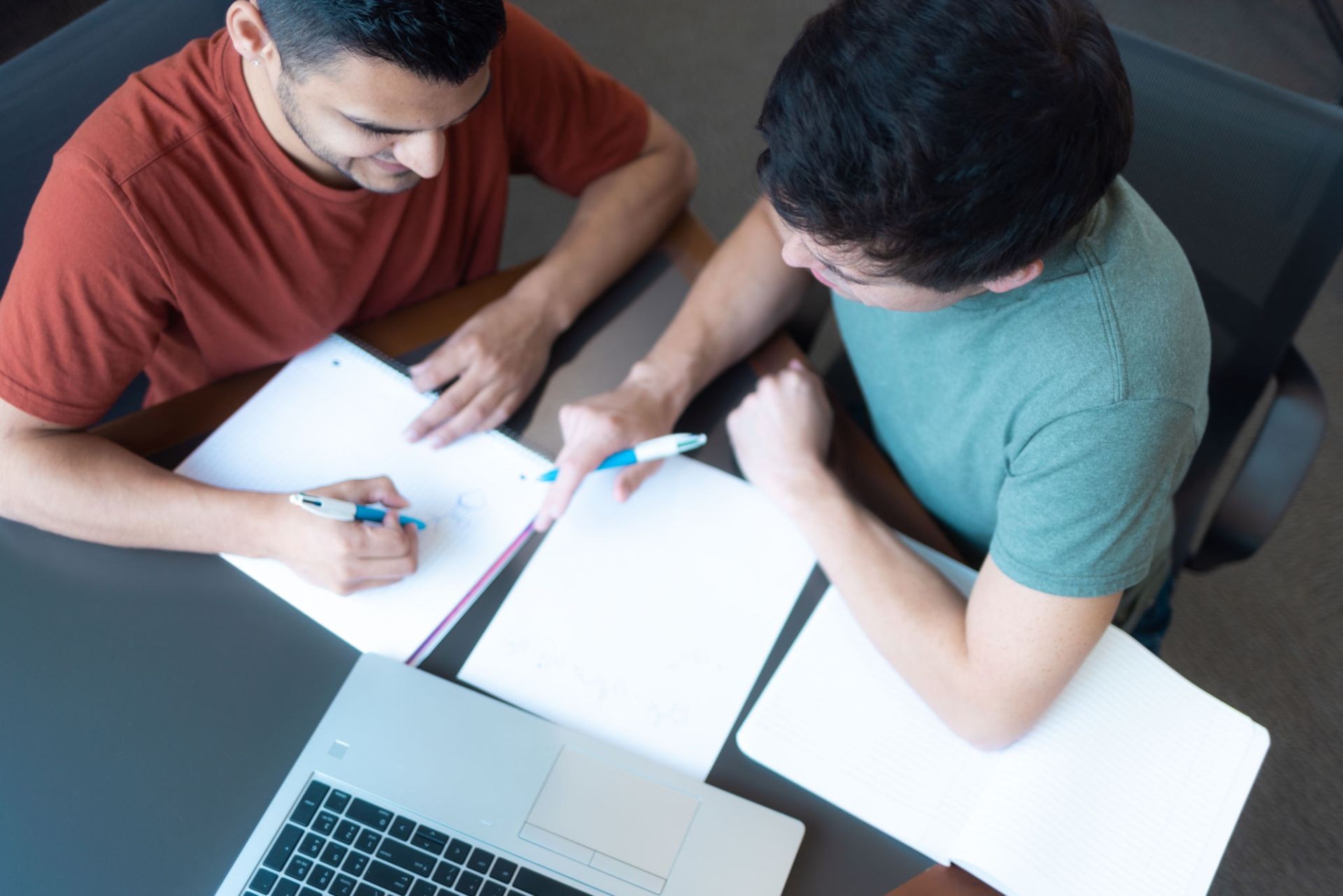 Two people sitting at a table with notebooks and a laptop, discussing and writing with pens.